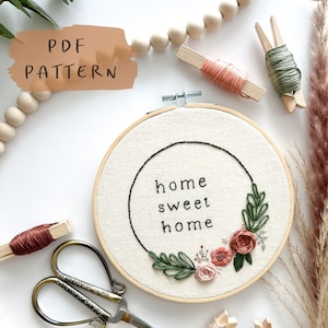 May include: A white embroidery hoop with the phrase "home sweet home" stitched in black thread. The hoop is surrounded by green leaves and pink flowers, and there are various embroidery supplies scattered around it.