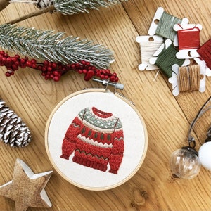 Puede incluir: Dise&ntilde;o de su&eacute;ter de Navidad bordado en un aro de bordado de madera. El su&eacute;ter es rojo, blanco y verde con un patr&oacute;n de copo de nieve.