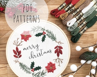 May include: A white embroidery hoop featuring a Christmas wreath design. The wreath is made of green leaves, red poinsettias, and holly. The text "merry christmas" is embroidered in black thread.