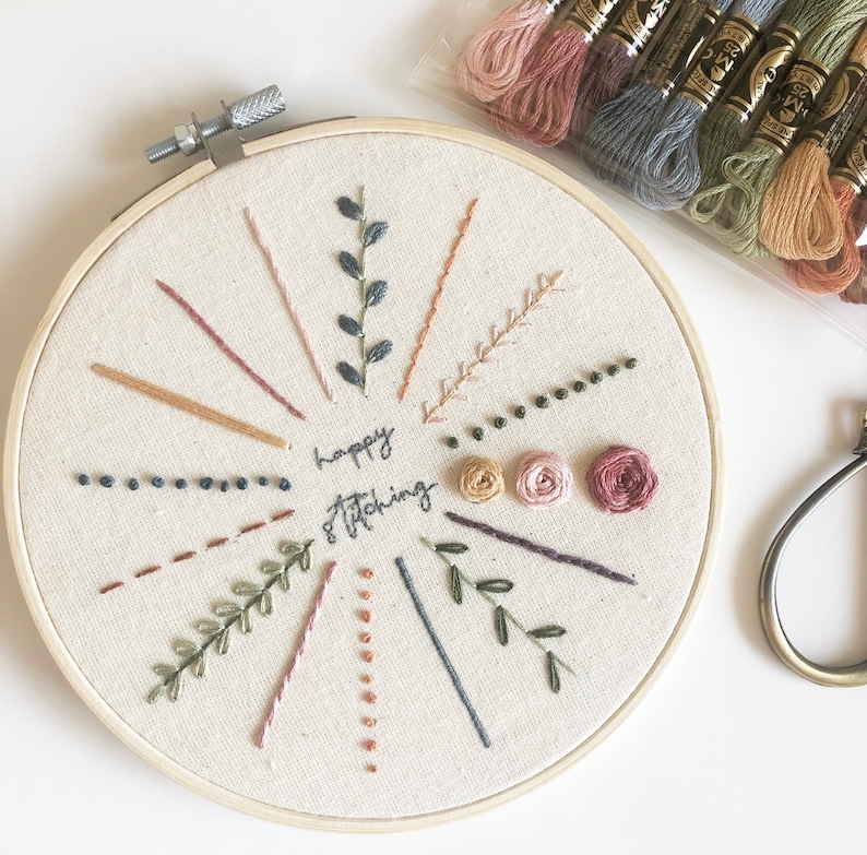 May include: A white embroidery hoop with a beige fabric background. The fabric is embroidered with various stitches, including lines, dots, and leaves. The text "happy stitching" is embroidered in the center of the hoop.