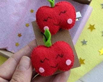 Puede incluir: Dos pinzas para el pelo de fieltro rojo con forma de cerezas con caras sonrientes y hojas verdes. Cada cereza tiene un punto blanco a cada lado. Las pinzas están sujetas a una tarjeta marrón.