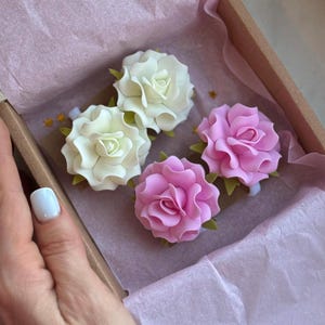 May include: Four artificial rose flowers in a box. Two are white, and two are pink. The flowers have green leaves and are arranged on a light pink surface. The box is brown and lined with pink tissue paper.