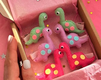 May include: A set of five felt dinosaur hair clips in a pink gift box. The dinosaurs are green, pink, and red with white polka dots.