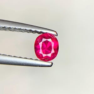 Könnte beinhalten: Ein oval geschliffener Rubin, gehalten von einer Pinzette. Der Rubin hat eine tiefe, rote Farbe, mit inneren Facetten, die ein Sternmuster erzeugen. Der Edelstein befindet sich vor einem schlichten, weißen Hintergrund.