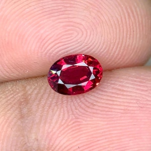 May include: A deep red oval gemstone with facets, set against a pink background.