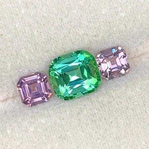 Pode incluir: Três pedras preciosas facetadas em tons de verde e roxo. A gema central é verde vibrante, ladeada por duas gemas roxas menores. Cada gema tem um corte quadrado com múltiplas facetas, criando um efeito brilhante.