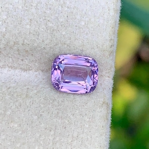 May include: A light purple, rectangular-cut gemstone with a square shape and rounded corners. The gem's internal facets reflect light. The stone is set against a light-coloured background.