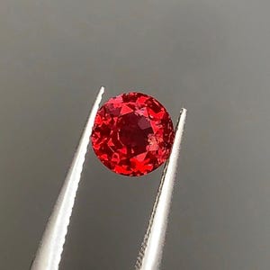 May include: A faceted, round, brilliant red gemstone is held by silver tweezers against a gray background. The gemstone's facets reflect light, creating a sparkling appearance. The tweezers are positioned diagonally, showcasing the gem's shape and color.
