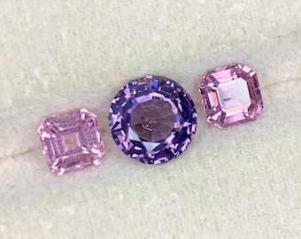 Può includere: Tre pietre preziose: una gemma viola rotonda e sfaccettata e due gemme rosa a taglio quadrato. Le gemme sono esposte su una superficie bianca e strutturata. Le gemme sono probabilmente destinate alla creazione di gioielli o al collezionismo.