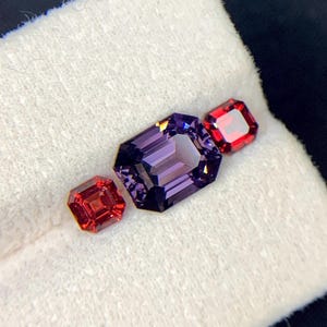 May include: Three gemstones on a white background. A purple gemstone is in the center, and two red gemstones are on either side of it.