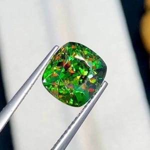 May include: A vibrant, cushion-cut gemstone held by silver tweezers. The gem features a mosaic of bright green, with flashes of red and orange. The facets catch the light, creating a sparkling effect, showcasing its unique color and cut.