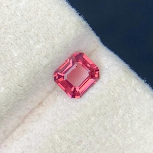 May include: A faceted, rectangular-cut, pink gemstone. The gem has a central rectangular facet and is surrounded by a series of smaller facets. The stone is set against a white, textured background.