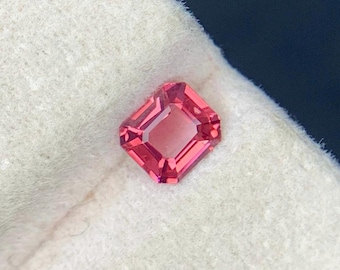 Op de afbeelding: Een roze edelsteen met rechthoekige slijpvorm en facetten. De steen heeft een centrale rechthoekige facet en is omgeven door kleinere facetten. Het is geplaatst tegen een witte, getextureerde achtergrond.