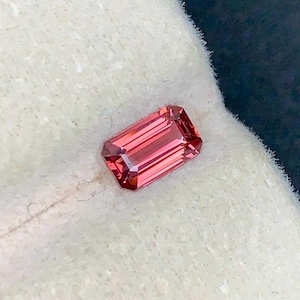 May include: A faceted, rectangular gemstone in a deep reddish-pink colour. The gem has a series of parallel lines visible within its structure, and is set against a white, textured background. The cut of the gem is an emerald cut.