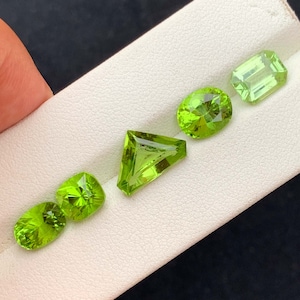 Op de afbeelding: Vijf groene peridoot edelstenen in verschillende vormen en maten op een witte displaykaart.