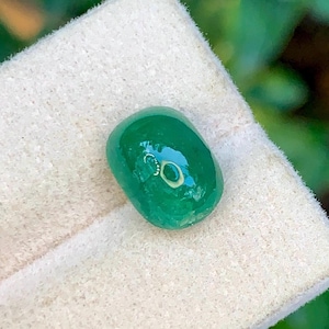 Op de afbeelding: Een gepolijste, ovaalvormige, smaragdgroene edelsteen. De edelsteen is glad en reflecterend, met een lichte glans. Hij ligt op een witte, getextureerde achtergrond, wat duidt op een presentatie. De levendige kleur en vorm van de edelsteen zijn de focus.