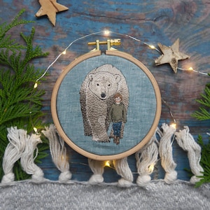 May include: Embroidered hoop art featuring a polar bear and a person standing next to it. The hoop is made of wood and has a blue linen fabric background.