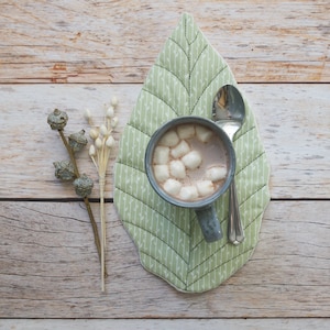 Può includere: Un sottobicchiere verde a forma di foglia con un motivo bianco e verde. Una tazza blu con cioccolata calda e marshmallow è appoggiata sul sottobicchiere. Un cucchiaio d'argento è accanto alla tazza.