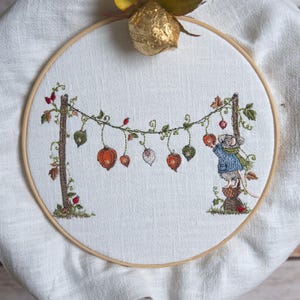 May include: An embroidery hoop containing white fabric with a stitched scene of a mouse in a blue coat standing on a stump, reaching for orange physalis lanterns hanging from a vine strung between two brown wooden posts with green leaves.