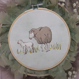 Könnte beinhalten: Gesticktes Bild in einem Holzrahmen, das ein Schaf und ein Lamm auf einer Wiese zeigt. Das Design ist vor einem hellgrünen Stoff angeordnet. Der Rahmen ist von einem Kranz aus grünen Federn umgeben.
