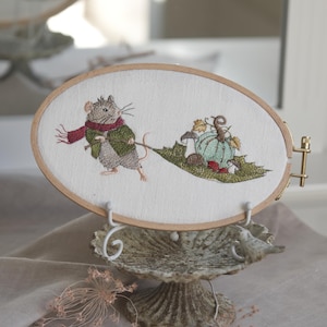May include: An oval wooden embroidery hoop containing white fabric with a detailed thread-stitched illustration of a gray mouse wearing a green jacket and red scarf, pulling a large green leaf carrying a light blue pumpkin and small mushrooms.