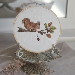 May include: A circular wooden embroidery hoop containing white fabric with a detailed thread-stitched design of a brown squirrel perched on a tree branch. The branch features green oak leaves and two acorns, with one additional acorn hanging below.