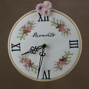 May include: White embroidered clock with black hands and Roman numerals. The clock face has the word "Moments" embroidered in cursive script and floral designs around the edge. The clock is decorated with pink flowers.