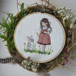 May include: Embroidered hoop art featuring a girl in a pink dress and a white bunny rabbit. The girl is holding a flower and the bunny is looking at her. The hoop is surrounded by dried flowers and grasses.