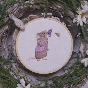 Könnte beinhalten: Gesticktes Bild im Stickrahmen mit einer braunen Maus in lila Weste und Schal, die einen Lavendelzweig hält. Eine gelb-schwarze Biene schwebt in der Nähe. Das Kunstwerk ist vor weißem Stoff, umrahmt von einem Holzreifen und einem dekorativen Kranz.