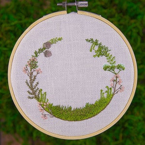 May include: A circular embroidery hoop with a white linen fabric background. The fabric is embroidered with a green wreath of leaves and flowers. The wreath is made up of small pink flowers, green leaves, and a few brown seed pods.