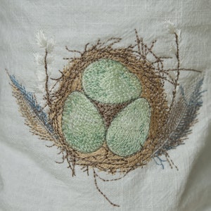 May include: Embroidered design of a bird's nest with three green eggs on a white fabric background.