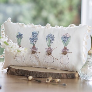 Könnte beinhalten: Ein weißes Kissen mit gestickten Hyazinthen in Blau, Grün und Rosa in Glasvasen, die mit Schnur gebunden sind. Das Kissen hat einen Fransenrand und liegt auf einer Holzoberfläche.