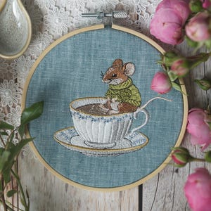 Könnte beinhalten: Stickerei in einem runden Rahmen mit einer braunen Maus, die einen grünen Pullover trägt und in einer weißen Teetasse mit brauner Flüssigkeit sitzt. Die Teetasse steht auf einem weißen Unterteller mit goldener Verzierung. Der Rahmen befindet sich auf einer Holzoberfläche mit rosa Blumen.