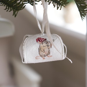 Könnte beinhalten: Gestickter Weihnachtsschmuck mit einer Maus mit rotem und weißem Hut. Das weiße Ornament hat ein gesticktes Bild der Maus. Ein weißes Band ist zum Aufhängen am Weihnachtsbaum befestigt.