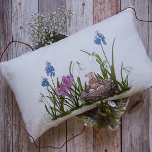 May include: A rectangular white cushion with an embroidered spring scene. The design features a bird in a nest with eggs, surrounded by purple crocuses, blue grape hyacinths, and white snowdrops. Green stems and leaves complete the floral arrangement.