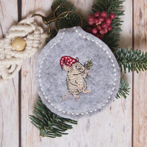 May include: A grey felt keyring with a stitched white border and a bronze-coloured clasp. The keyring features an embroidered mouse wearing a red and white polka-dot hat and holding a sprig of greenery. Macrame and greenery are also visible.