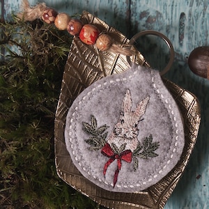 May include: A gray felt keychain with an embroidered image of a brown rabbit wearing a red bow and surrounded by green leaves. The keychain is attached to a gold metal leaf-shaped charm.
