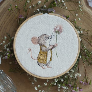 May include: Embroidered hoop art featuring a brown mouse with a yellow waistcoat holding a pink flower. The mouse is embroidered on a white fabric background.