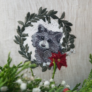 Op de afbeelding: Borduurwerk van een wasbeer met een rode kerststerbloem op een witte linnen stoffen achtergrond. De wasbeer wordt omringd door een groene krans.