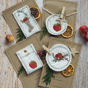 Könnte beinhalten: Eine Sammlung handgefertigter Weihnachtskarten und Geschenkanhänger. Die braunen Karten haben genähte Ränder und gestickte Designs von Strümpfen, Äpfeln und Erdbeeren. Getrocknete Orangenscheiben und grüne Zweige sorgen für festliche Akzente.