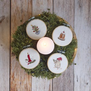 Peut inclure: Une composition festive avec cinq cerceaux brodés ornés de motifs de Noël. Les motifs incluent un sapin, un pudding, une bougie et une souris portant un chapeau. Une bougie allumée est au centre, entourée de verdure, créant une ambiance chaleureuse.