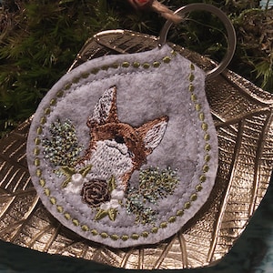 May include: A gray felt keychain featuring an embroidered fox head with brown and white details, surrounded by floral embroidery in green, white, and brown. The keychain has a silver metal ring and a decorative green stitched border.