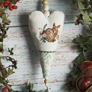 Könnte beinhalten: Ein weißer herzförmiger Stoffschmuck mit einem grün-weißen Blumenmuster und einem gestickten Bild eines Rehs mit Blumen. Der Schmuck hängt an einer Schnur aus Holzperlen.