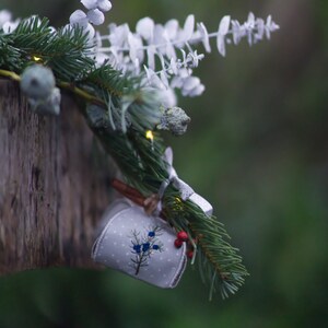 May include: A white fabric ornament with a blue and green floral design hangs from a pine branch with white berries. The ornament is tied with twine and a white ribbon.