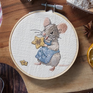 Könnte beinhalten: Stickerei in einem Holzreifen mit einer braunen Maus, die einen gelben Stern hält. Die Maus trägt eine blaue und weiße Schürze. Der Reifen ist aus Holz und hat eine natürliche Oberfläche.