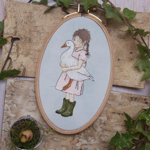 May include: An embroidered artwork in an oval wooden frame. The artwork depicts a child in a pink dress and green boots holding a white goose. The background is a light blue color. The embroidery is detailed and colorful.