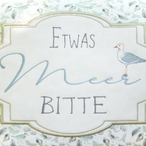 May include: A white embroidered pillow with a green border and a seagull design. The pillow features the text "Etwas Meer Bitte" in German.