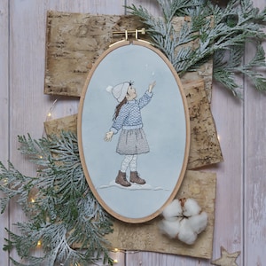 May include: An oval embroidery hoop depicts a girl in a winter scene, wearing a blue sweater, grey skirt, and white hat, reaching up towards falling snow. The artwork is set against a light blue background, with a wooden frame. Birch bark, greenery, and a cotton ball are also present.