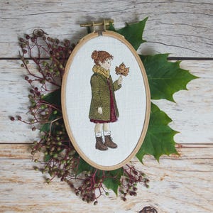 May include: An embroidered artwork in an oval wooden hoop. The artwork depicts a child in a green coat and scarf holding a fall leaf. The embroidery is set against a white background. The hoop is surrounded by greenery.