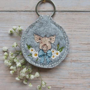 May include: A gray felt keychain with a stitched design of a pig wearing a blue bow, surrounded by embroidered daisies. The keychain has a silver metal ring attached at the top. White flowers are arranged on the left side of the image.